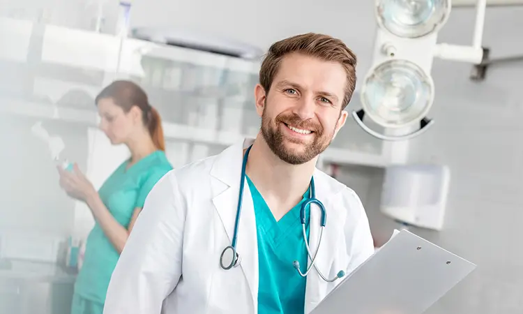 A man in a lab coat smiles and holds a clipboard, with a stethoscope hanging from his neck. In the background, a woman works in a medical setting.